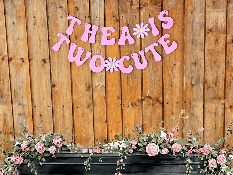 Pu&ograve; includere: Striscione rosa con il testo "Thea is Two Cute" con fiori bianchi tra le parole. Lo striscione &egrave; appeso a una parete di legno con una ghirlanda di fiori sotto.