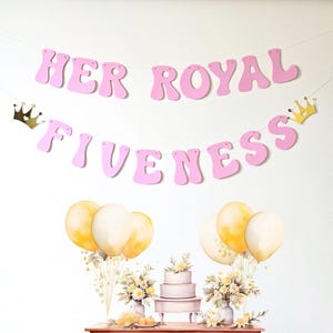 May include: Pink banner with the text "Her Royal Fiveness" in a whimsical font. The banner is decorated with gold crowns and is strung with white thread. There are also gold balloons and a white cake on a table in the background.