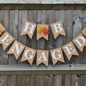Puede incluir: Una pancarta marrón con letras blancas que deletrean "ENGAGED" y "H & B". La pancarta está decorada con hojas de otoño. La pancarta está colgada frente a una valla de madera.