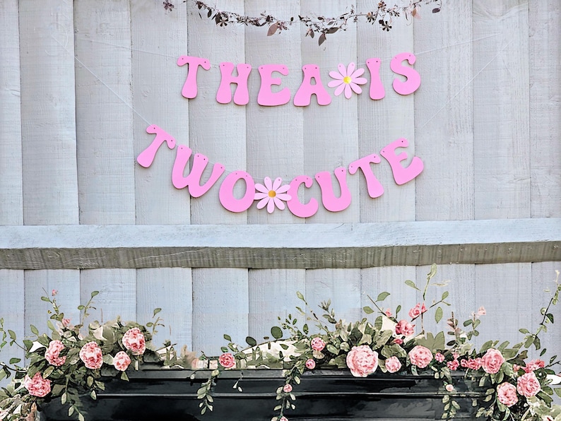 Pu&ograve; includere: Striscione rosa con il testo "Thea is Two Cute" con margherite bianche tra le parole. Lo striscione &egrave; appeso a una recinzione di legno bianca. Una ghirlanda di fiori rosa &egrave; davanti alla recinzione.