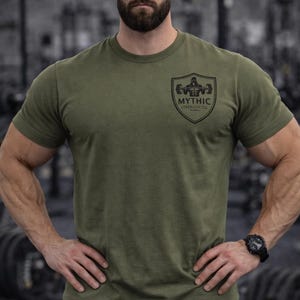 May include: Olive green t-shirt with a black logo that reads "Mythic Strength Co." The shirt is short-sleeved and has a crew neck. The background is a gym setting with workout equipment.