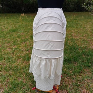 May include: White cotton petticoat with a ruffled bottom and multiple layers. The petticoat is on a dress form and is in a grassy area.