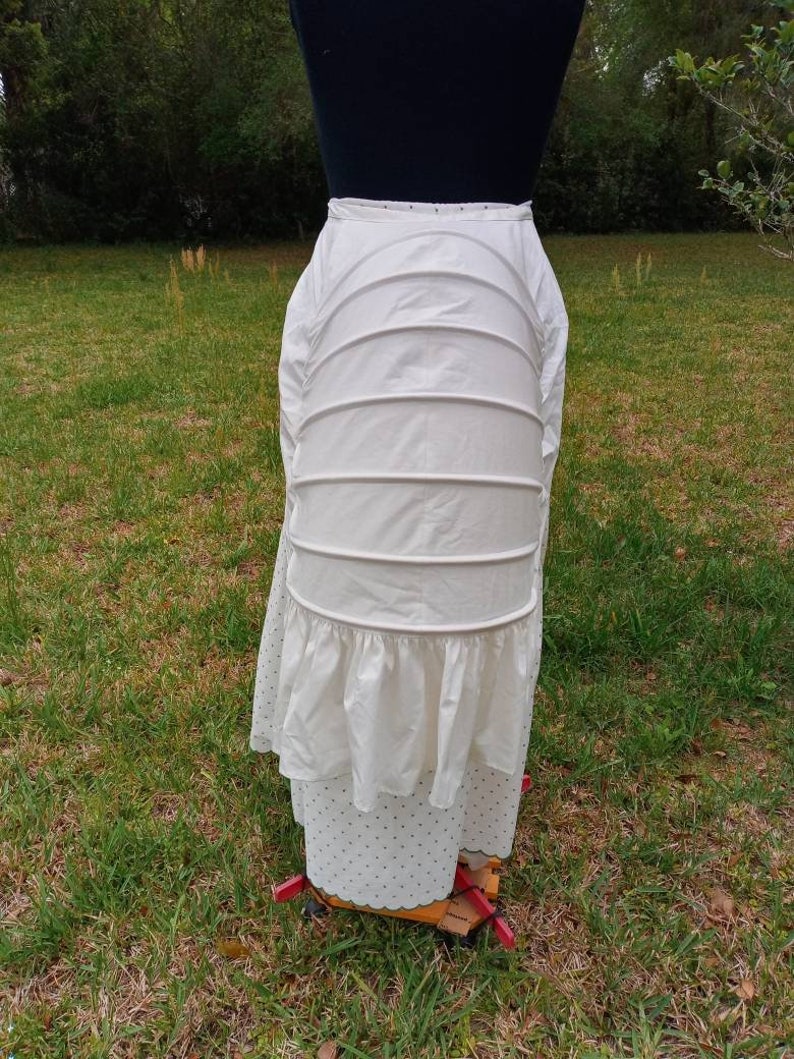 May include: White cotton petticoat with a ruffled bottom and multiple layers. The petticoat is on a dress form and is in a grassy area.