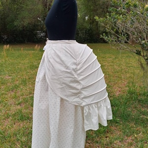 May include: A white cotton bustle with a ruffled edge. The bustle is worn over a white skirt with black polka dots.