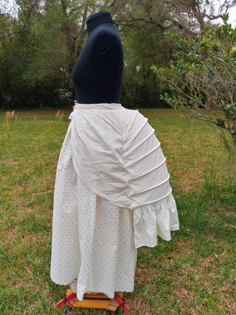 May include: A white cotton bustle with a ruffled edge. The bustle is worn over a white skirt with black polka dots.
