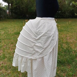 May include: A white cotton bustle with a ruffled edge. The bustle is worn over a white skirt with black polka dots.