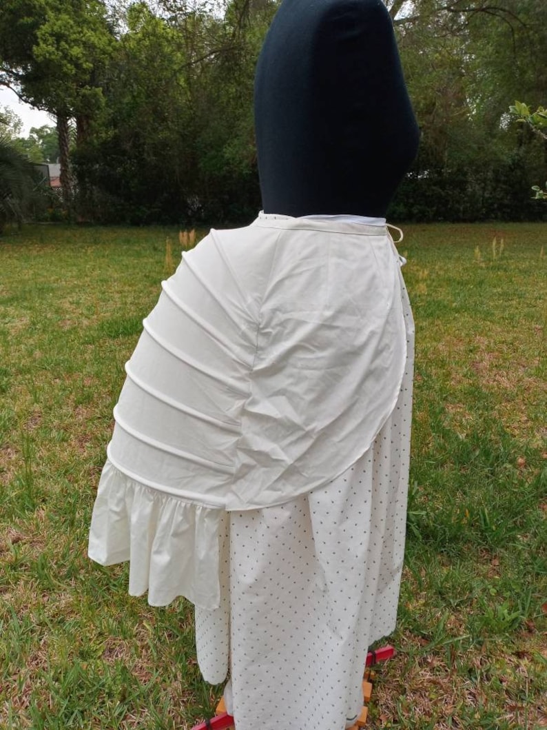 May include: A white cotton bustle with a ruffled edge. The bustle is worn over a white skirt with black polka dots.