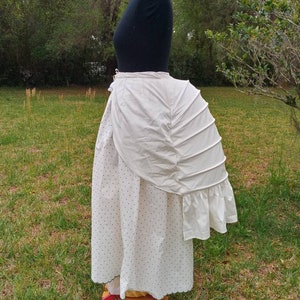 May include: A white cotton bustle with a ruffled edge and pleats. The bustle is worn over a white skirt with black polka dots.