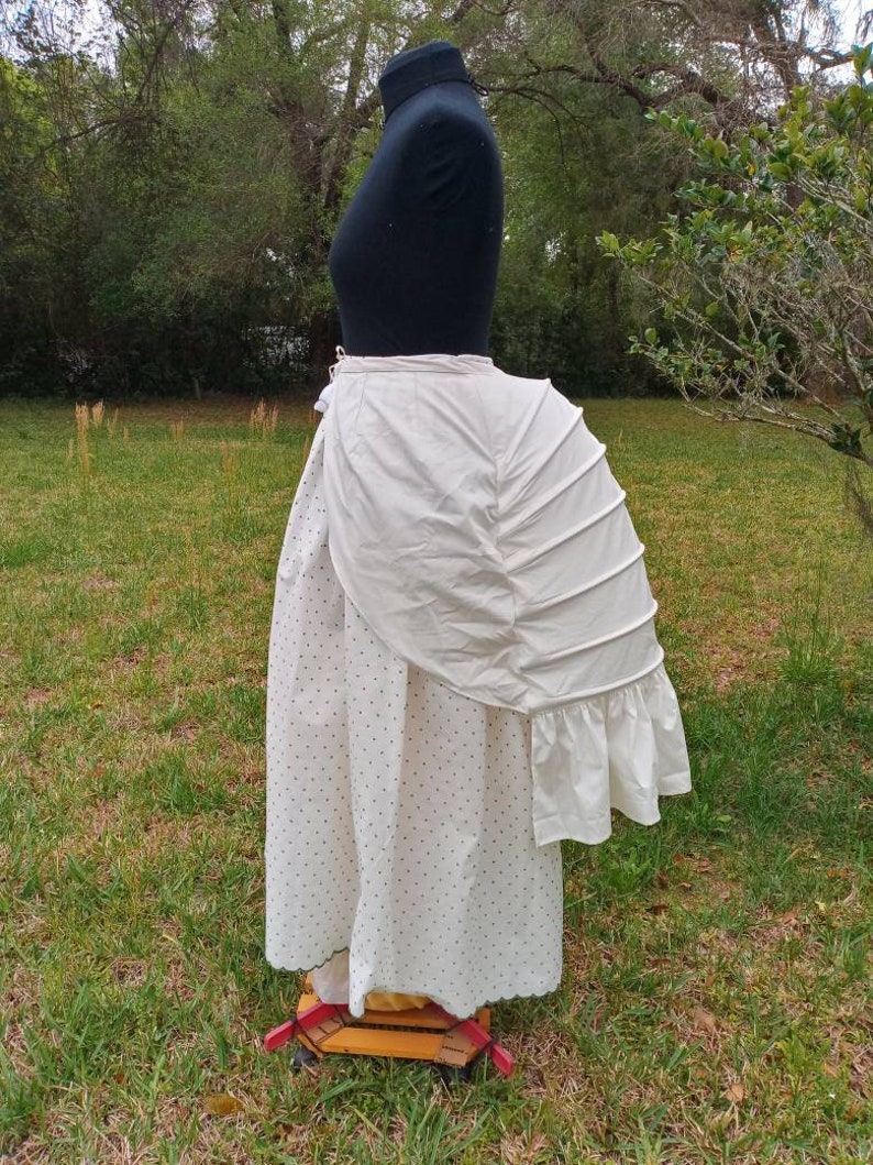 May include: A white cotton bustle with a ruffled edge and pleats. The bustle is worn over a white skirt with black polka dots.
