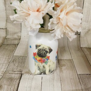 May include: A white ceramic jar decorated with a floral design and a pug wearing a flower garland. The jar is filled with a bouquet of pale pink peonies.