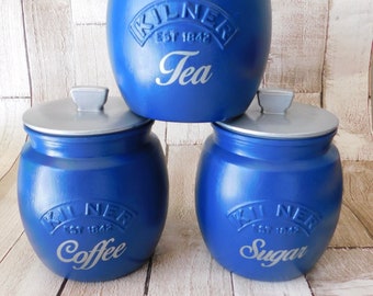 Op de afbeelding: Drie blauwe keramische potten met zilveren deksels. De potten zijn gelabeld met "Thee", "Koffie" en "Suiker" met het Kilner-logo en "Est 1842".