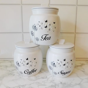 May include: Three white ceramic canisters with black and grey dandelion and butterfly designs. The canisters are labelled "Tea", "Coffee", and "Sugar".