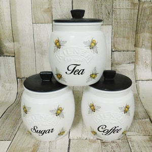 May include: Three white ceramic canisters with black lids and bee designs. The canisters are labelled "Tea", "Sugar", and "Coffee".