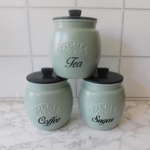 May include: Set of three green ceramic canisters with black lids and black lettering. The canisters are labelled "Tea", "Coffee", and "Sugar".