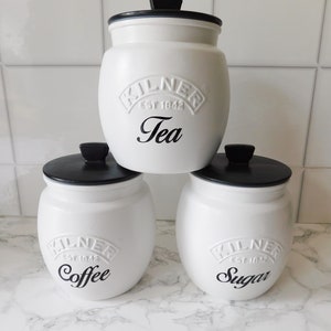 May include: Three white ceramic canisters with black lids and black lettering. The canisters are labelled "Tea", "Coffee", and "Sugar".