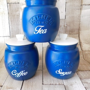 May include: Three blue ceramic canisters with white lids and white lettering. The canisters are labelled "Tea", "Coffee", and "Sugar".