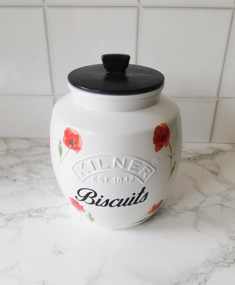 May include: White ceramic biscuit jar with a black lid and handle. The jar is decorated with red poppy flowers and the word "Biscuits" in black script. The jar has the "Kilner Est 1842" logo.