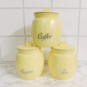 May include: Three pale yellow ceramic storage canisters with matching lids. Each canister features the embossed Kilner logo and the text Coffee, Sugar, or Tea in a script font. The canisters are arranged in a pyramid formation on a surface.
