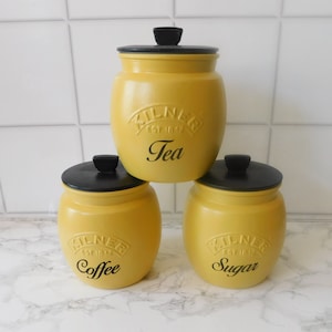 May include: Three yellow ceramic canisters with black lids and black lettering. The canisters are labelled "Tea", "Coffee", and "Sugar".
