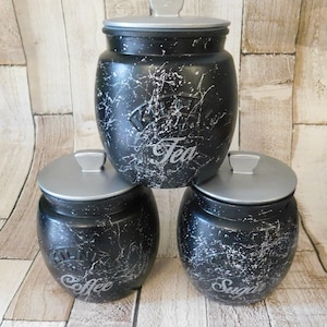 May include: Three black ceramic canisters with silver lids and white splatter paint. The canisters are labelled "Coffee", "Tea", and "Sugar".