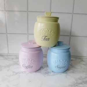 May include: Three ceramic canisters with lids, labelled "Tea", "Coffee", and "Sugar". The canisters are pastel yellow, pink, and blue.