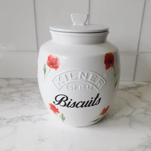May include: White ceramic biscuit jar with a lid, featuring red poppy flower designs. The jar has the word "Biscuits" in black script and the embossed "Kilner" logo.