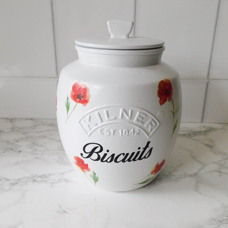 May include: White ceramic biscuit jar with a lid, featuring red poppy flower designs. The jar has the word "Biscuits" in black script and the embossed "Kilner" logo.