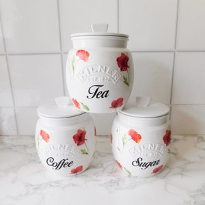May include: Three white ceramic canisters with red poppy floral designs and black lettering. The canisters are labelled "Tea", "Coffee", and "Sugar".
