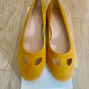 May include: A pair of mustard yellow suede ballet flats with a rounded toe and two oval cutouts. The shoes are displayed on a white box, set against a light wood grain background. The interior of the shoe is a light tan color.
