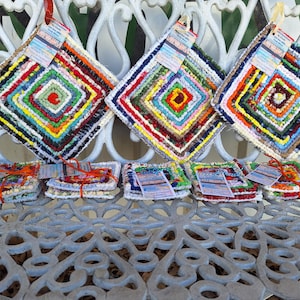 May include: A collection of handmade, square, colorful crochet potholders and coasters. The potholders are diamond-shaped with a variety of colors, including red, green, blue, and yellow. The coasters are square and stacked in small bundles.