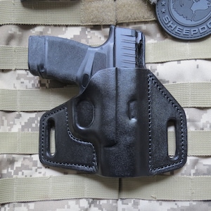 May include: Black leather holster for a handgun. The holster has a belt loop and a snap closure. The holster is on a camouflage background.