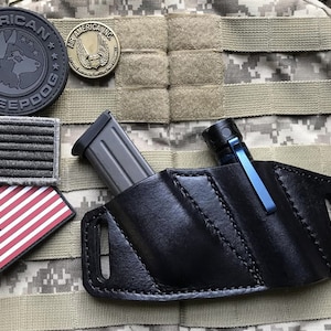 May include: A black leather magazine and flashlight pouch with a magazine and flashlight inside. The pouch is attached to a camouflage MOLLE panel with American flag patches.