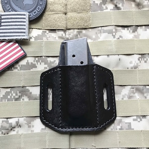 May include: A black leather magazine pouch with a metal magazine inside. The pouch is attached to a camouflage tactical vest.