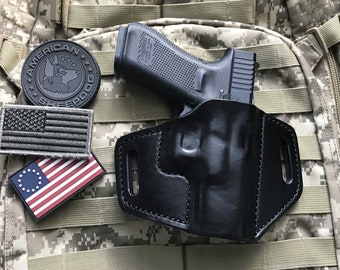 Puede incluir: Funda de cuero negra para una pistola, con una pistola negra en su interior. La funda está sobre un fondo de camuflaje con parches de la bandera estadounidense.