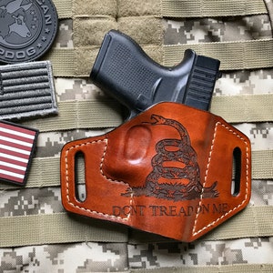 May include: A brown leather holster with a snake and the text "Don't Tread On Me" engraved on it. A black handgun is in the holster. The holster is on a camouflage background.