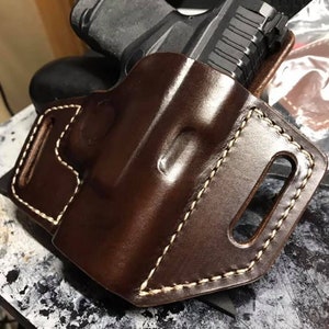 Puede incluir: Una funda de cuero marr&oacute;n con costuras blancas para una pistola. La funda tiene un bucle para el cintur&oacute;n y un fondo abierto.