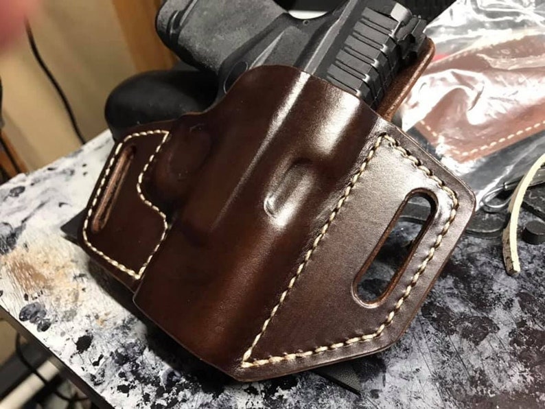 Puede incluir: Una funda de cuero marr&oacute;n con costuras blancas para una pistola. La funda tiene un bucle para el cintur&oacute;n y un fondo abierto.