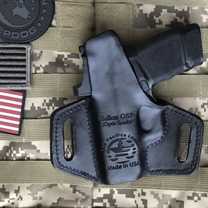 Puede incluir: Funda de cuero negra con una pistola negra en su interior. La funda tiene el texto "Hellcat OSP (Optic Ready)" y "New Pacific Leather Made in USA" grabado en relieve.