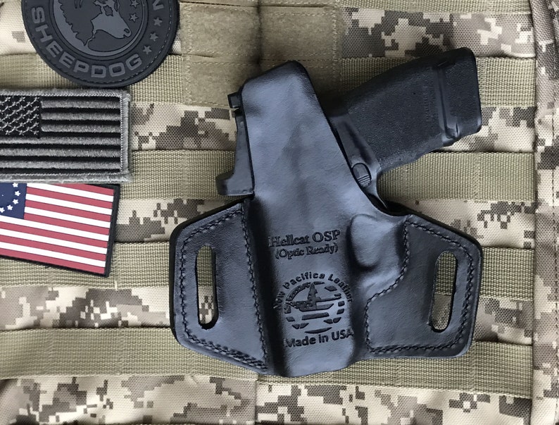Puede incluir: Funda de cuero negra con una pistola negra en su interior. La funda tiene el texto "Hellcat OSP (Optic Ready)" y "New Pacific Leather Made in USA" grabado en relieve.