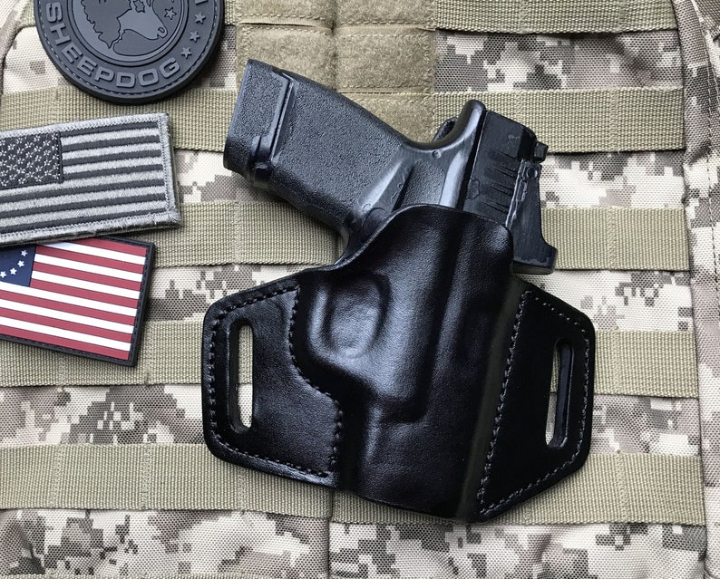 Puede incluir: Una funda de cuero negra con una pistola negra en su interior. La funda tiene un bucle para el cintur&oacute;n y un cierre a presi&oacute;n. La pistola es una pistola semiautom&aacute;tica con un marco de pol&iacute;mero negro y un deslizador de acero inoxidable.