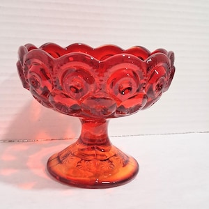 May include: A vibrant red glass compote dish featuring a scalloped rim and a repeating moon and star pattern embossed around the bowl and the circular base. The pedestal design elevates the bowl, which has a translucent finish and a glossy texture.