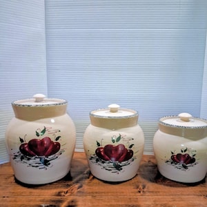 May include: Set of three ceramic canisters with lids. The canisters are off-white with a cream-colored rim and lid. Each canister features a hand-painted design of red flowers with green leaves and black accents. The canisters are of varying sizes.