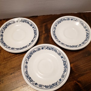 May include: Three white saucers with a blue floral and leaf border are arranged on a dark wood surface. The saucers have a central indentation for a cup and a decorative edge.