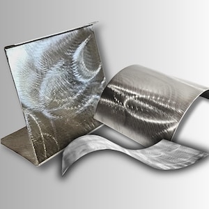 May include: Abstract metal art sculpture with a brushed finish. The sculpture features a square piece, a curved piece, and a flowing, wave-like piece. The metal has a silver color and a textured appearance, creating a modern aesthetic.