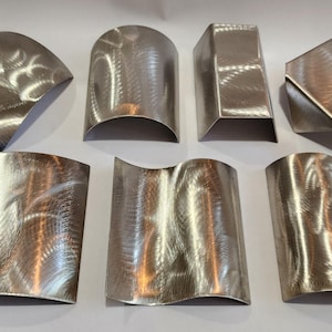 May include: A collection of seven abstract metal sculptures with a brushed finish. The shapes include curved, angular, and geometric forms, showcasing a variety of designs. The sculptures are displayed on a white surface.
