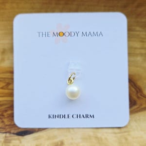 Peut inclure: Une carte blanche aux coins arrondis affiche le texte "THE MOODY MAMA" et "KINDLE CHARM". Un petit charme doré avec une perle blanche est suspendu à un clip en plastique transparent. Le fond est une surface en bois.