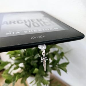 May include: A silver sword bookmark attached to a black Kindle e-reader with the text "Archer's Voice" and "MIA SHERIDAN" on the screen. The Kindle is resting on a wooden surface with a potted plant in the foreground.