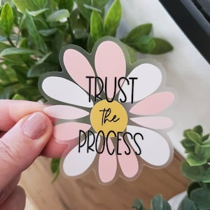 May include: A clear sticker featuring a pink and white daisy design. The daisy has a yellow centre and the text "TRUST the PROCESS" in black.