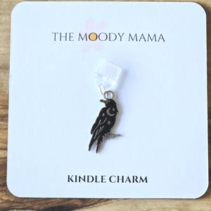 Peut inclure: Un charm en forme de corbeau noir avec un croissant de lune et des étoiles, attaché à un fermoir en plastique transparent. Le charm est sur une carte blanche avec le texte "THE MOODY MAMA" et "KINDLE CHARM".