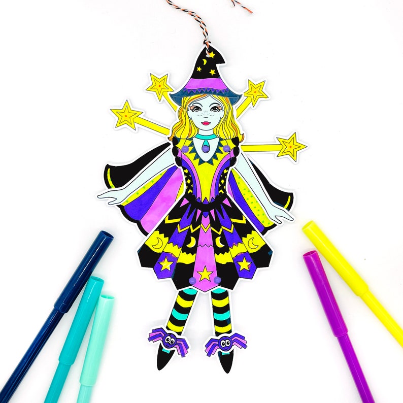 May include: A paper craft witch doll with a purple and yellow dress, black cape, and star-shaped wands. The doll is ready to be colored with markers.